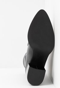 Botte noire à talon haut avec un bout pointu et une texture lisse. Semelle côtelée et finition brillante. Bloc talon avec des détails subtils.