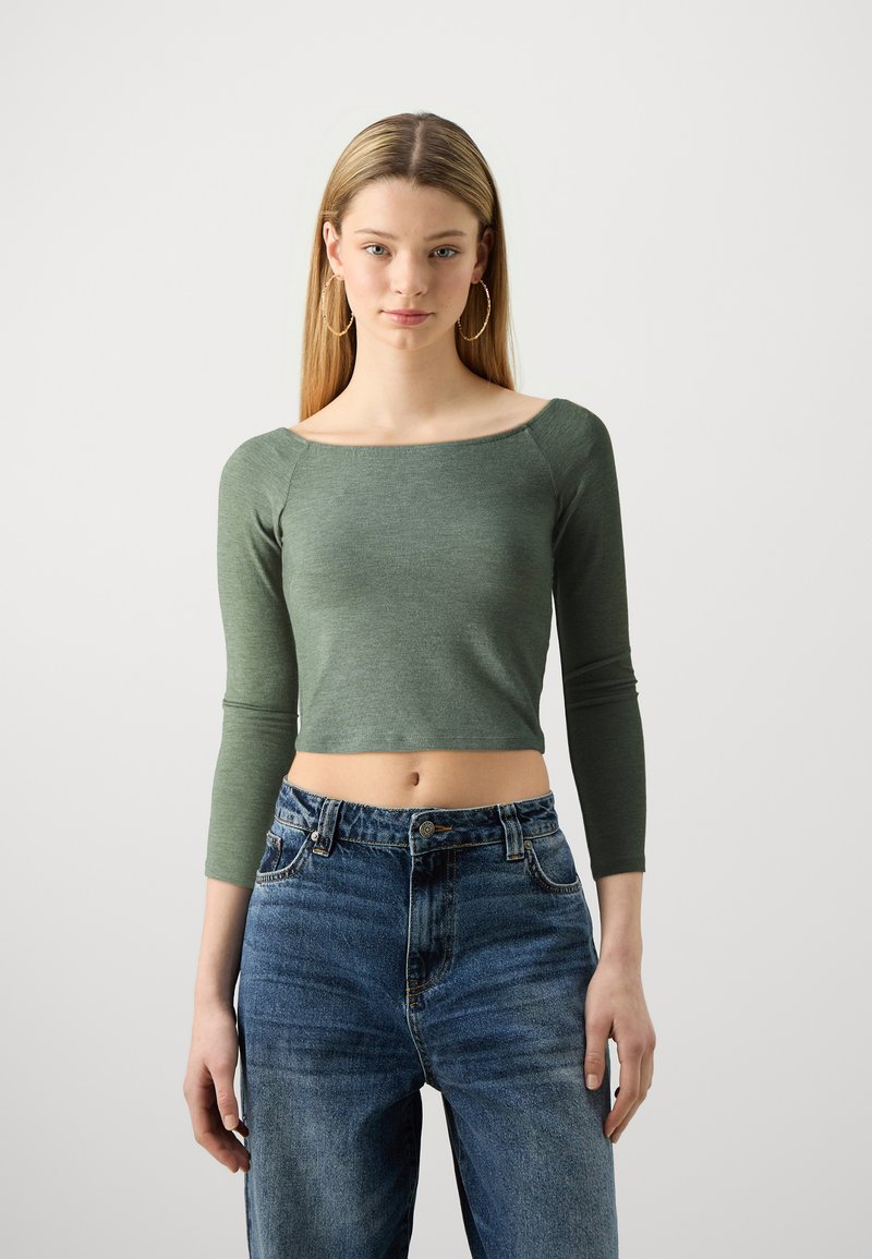 Zöld, testhez simuló, hosszú ujjú crop top, négyzet alakú kivágással; magas derekú, megkopott kék farmernadrággal párosítva, amelyen varrási részletek találhatók.