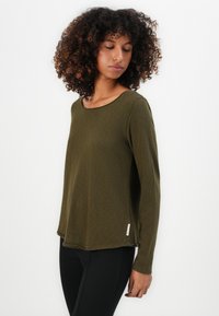 Donna con capelli ricci che indossa una maglietta verde oliva a maniche lunghe e pantaloni neri, che guarda in basso contro uno sfondo bianco uniforme.