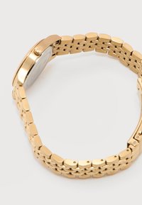 Χρυσό μεταλλικό ρολόι με συνδεδεμένο βραχιόλι και κλειστό κούμπωμα, εμφανιζόμενο από το πλάι σε λευκό φόντο.