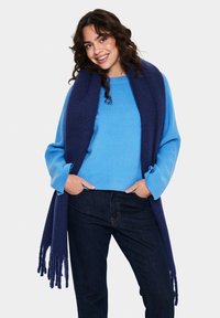 Blue knit sweater, dark denim jeans, and a long navy scarf with fringe details. The sweater features ribbed cuffs and a relaxed fit.