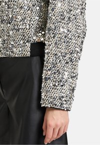 Paillettenjacket met een gestructureerde stof in zwart, wit en zilver; heeft een geribbelde zwarte manchette; gedragen over zwarte leren broek.