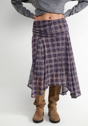 Spódnica w średniej długości fioletowa w kratę z marszczonym pasem, noszona z wysokimi brązowymi zamszowymi butami i krótkim szarym swetrem.