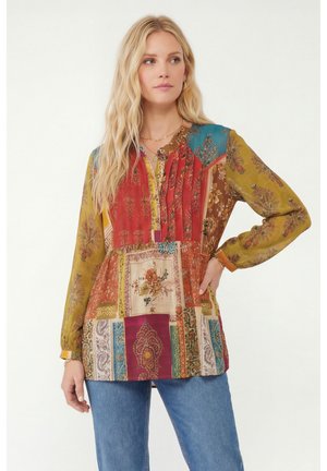Femme blonde portant un chemisier patchwork coloré avec des imprimés floraux et cachemire, associé à un jean bleu, debout devant un fond blanc.