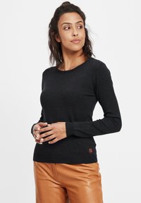 Zwarte lange mouwen trui met een ronde halslijn, geribbelde manchetten en een klein bruin label aan de zoom, gecombineerd met lichtbruine leren broeken.