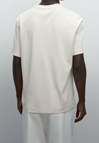 Man som bär en enkel ljusgrå kortärmad t-shirt och vita byxor, sedd bakifrån mot en neutral grå bakgrund.