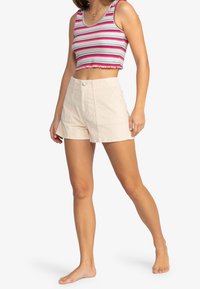 Débardeur court rayé en tissu rose, blanc et bleu ; associé à un short taille haute beige avec deux poches avant et une fermeture à bouton.