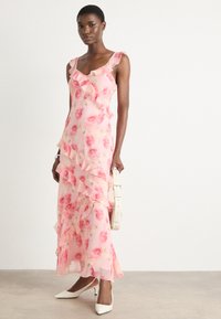 Long pink floral dress with ruffle details, made of lightweight fabric. Features slits and a fitted bodice. Model holds a white belt bag.