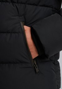 Giacca a vento nera con tasca con zip e hardware dorato, texture trapuntata visibile e design leggermente imbottito per l'isolamento.