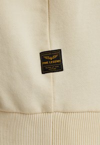 La tela de mezcla de algodón beige presenta un dobladillo acanalado texturizado y una etiqueta prominente en negro y dorado con "PME LEGEND" y "TESTED AT HIGH ALTITUDE."