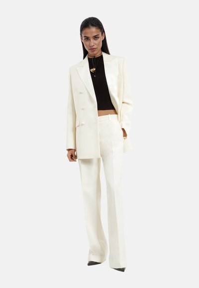 Completo color crema con giacca doppiopetto e pantaloni a gamba larga, abbinato a un crop top nero a coste; include una collana a forma di cuore dorata.