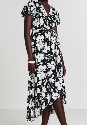 Schwarz-weißes, geblümtes Wickelkleid mit kurzen Ärmeln und asymmetrischem Saum, getragen von einer Person mit dunkler Haut, accessoriert mit weißen Armreifen.