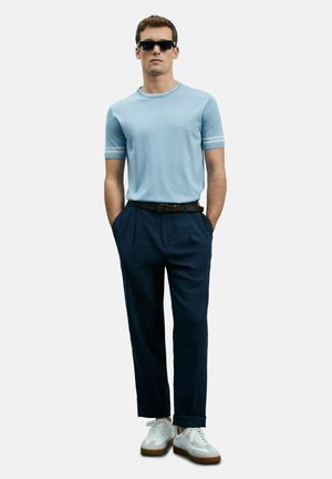 Hellblaues kurzärmliges Sweatshirt mit weißen Streifen an den Bündchen, kombiniert mit dunklen marineblauen Hosen und weißen Sneakers mit braunen Sohlen. Sonnenbrille inklusive.