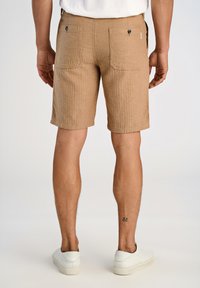 Bruna, texturerade shorts med rak skärning och bakfickor. Bärs med vita sneakers. Synlig tatuering på vänster ben.