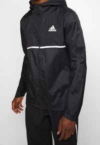 Man som bär en svart Adidas vindjacka med vitt logo och en reflekterande horisontell rand över bröstet.