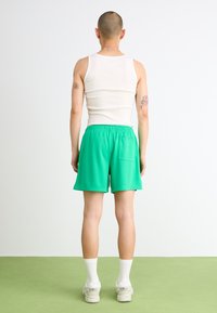 Şorturi atletice verzi din plasă cu un brâu elastic, buzunar posterior, purtate cu un maieu alb și ciorapi albi, stând pe un podea verde.