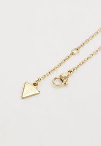 Necklace od zlatnog tona s trokutastim privjeskom ugraviranim s "GUESS?" i zatvaranjem na školjku.