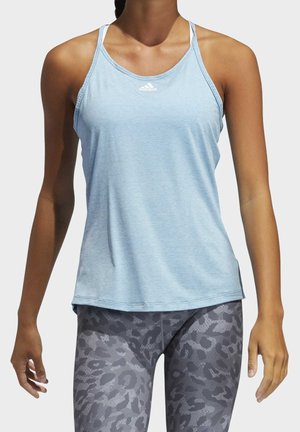 Vrouw draagt een lichtblauwe mouwloze Adidas tanktop en grijze legging met patroon, staand tegen een effen achtergrond.