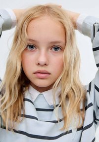 Pulover albastru deschis cu dungi închise, decolteu rotund și guler alb. Păr blond ondulat, lungime până la umeri, încadrează o expresie neutră.