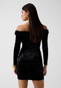 Kobieta z długimi, prostymi włosami, ubrana w czarną, aksamitną mini sukienkę z odkrytymi ramionami, złote kolczyki koła i złotą bransoletkę, zwrócona lekko w lewo.