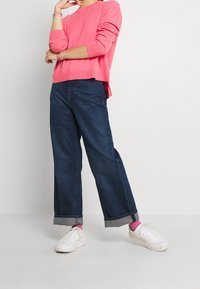 Hellrosa, langärmliger Pullover mit geripptem Saum, kombiniert mit dunkelblauen, weit geschnittenen Jeans, die am Saum umgeschlagen sind, und weißen Turnschuhen mit rosa Socken.