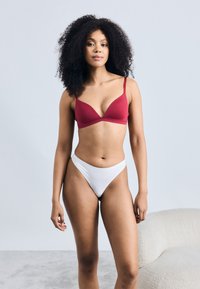 Κόκκινο τριγωνικό σουτιέν με λεπτές τιράντες που συνδυάζεται με λευκό ψηλόμεσο στρινγκ. Λείο υλικό, αόρατος σχεδιασμός και εφαρμοστό σχήμα.