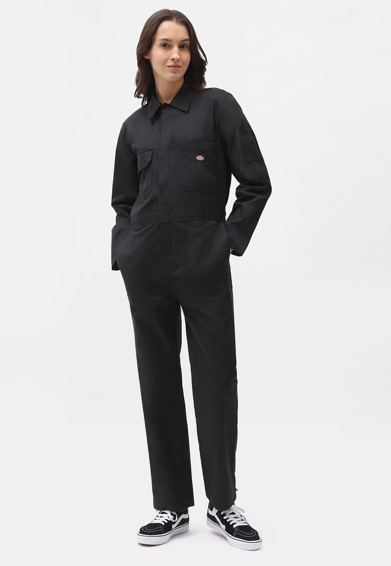 Zwarte overall gemaakt van duurzaam stof, met een kraag, een voorkantzak en een logo patch. Gemaakt in combinatie met zwart-witte sneakers.