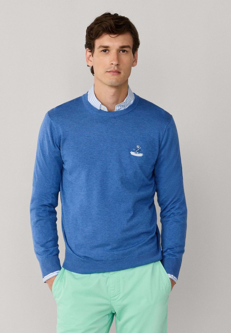 Pull bleu à col rond en tissu tricoté, avec un petit motif brodé sur la poitrine gauche et des poignets en côte, associé à un short vert menthe.