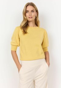Haut en tricot jaune avec un col rond, des manches courtes bouffantes et une texture douce, associé à un pantalon crème taille haute.