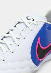 Λευκά δερμάτινα αθλητικά παπούτσια με λευκές κορδόνια, μπλε πλαϊνές πάνελ και μαύρο λογότυπο Nike περιγραμμένο σε ροζ, σε απλό φόντο.