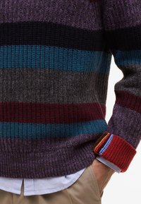 Chunky knit sweater featuring horizontal stripes in shades of purple, teal, grey, and burgundy. Ribbed cuffs with colorful accents.