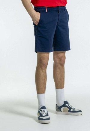 Shorts marine à devant plat, avec deux poches latérales et une ceinture colorée en couture. Associés à des baskets bleues et crème et des chaussettes blanches.