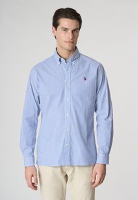 Camicia a righe blu e bianche con colletto, maniche lunghe e un piccolo logo rosso sul petto. Materiale in cotone, texture liscia.