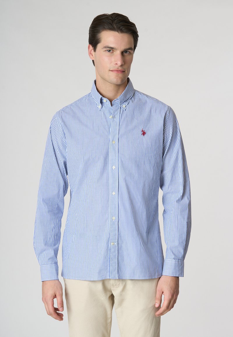 Camicia a righe blu e bianche con colletto, maniche lunghe e un piccolo logo rosso sul petto. Materiale in cotone, texture liscia.