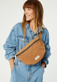 Sac banane en tissu marron avec fermeture éclair et étiquette logo, porté en bandoulière. Associé à une veste en denim bleu et à des salopettes bleu clair.