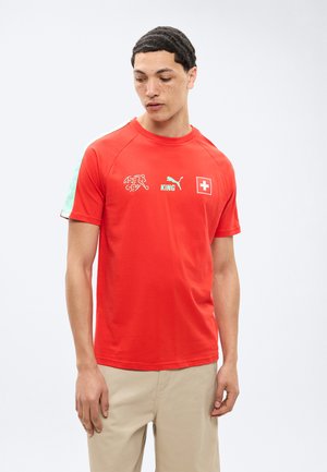 Νέος άνδρας που φοράει κόκκινο μπλουζάκι της ελβετικής ομάδας ποδοσφαίρου με το λογότυπο της Puma, το κείμενο "KING", και μπεζ παντελόνια, στέκεται μπροστά σε απλό λευκό φόντο.