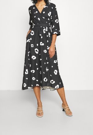 Vestito avvolgente nero con motivo floreale bianco, maniche lunghe, vita legata e orlo fluido che arriva sotto il ginocchio. Indossato con sandali con cinturini.