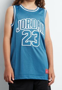 Modrý síťovaný basketbalový dres s bíl�ým lemováním, na kterém je velkými písmeny napsáno "JORDAN" a číslo "23", doplněný červenou logovou nášivkou.