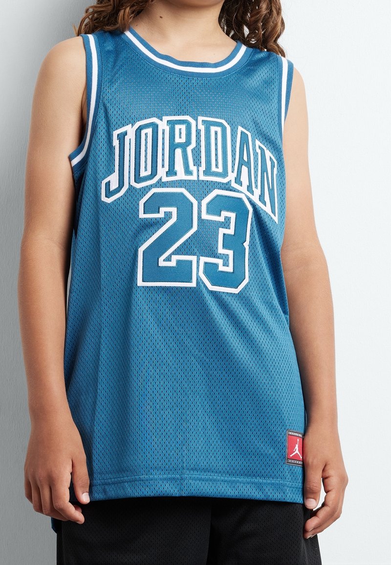 Modrý síťovaný basketbalový dres s bílým lemováním, na kterém je velkými písmeny napsáno "JORDAN" a číslo "23", doplněný červenou logovou nášivkou.