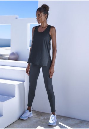 Dunkelgraues Sporttanktop und Leggings, die einen lockeren Schnitt und Seitenschlitze aufweisen. Weiße und blaue Sneakers runden das sportliche Outfit ab.