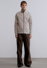 Μπεζ ζακέτα με φερμουάρ, γιακά που στέκεται, ριμπς στα μανίκια και στη μέση, σετ με φαρδιά καφέ παντελόνια. Το μοντέλο στέκεται σε ουδέτερο φόντο.