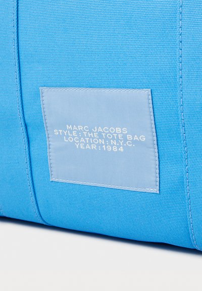 Borsa a tracolla in tela blu con un'etichetta azzurro chiaro contenente testo bianco che indica il marchio, lo stile, la posizione e l'anno. Esterno testurizzato con dettagli di cucitura.