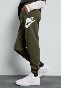 Olivově zelené tepláky s výrazným bílým logem Nike na noze, v kombinaci s bílými teniskami. Měkká tkanina s volným střihem.