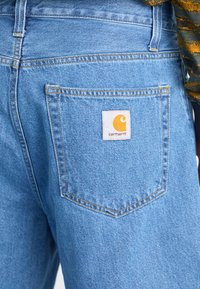 Blue denim jeans with a classic five-pocket design. Back pocket features a white fabric label with an orange logo. Visible stitching details.