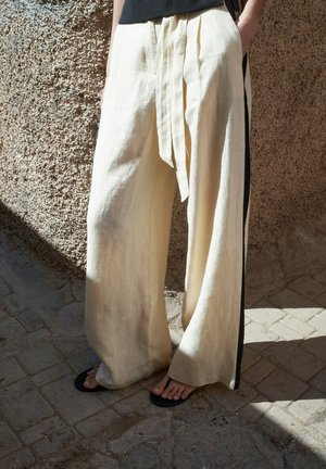 Personne portant un pantalon ample en lin beige clair avec une bande latérale noire, debout sur un pavé en pierre, pieds nus dans des sandales noires.