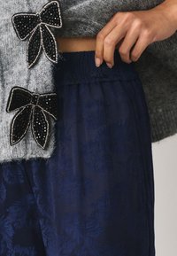 Pull gris en tricot avec des accents de nœud en perles noires ; pantalon bleu marine à taille élastique avec un motif subtil de feuilles. Texture douce et design superposé.