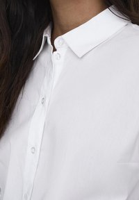 Nahaufnahme des Kragens eines weißen Hemdes mit Knöpfen und der oberen Brust, mit dunklen Haaren auf der linken Seite, die die glatte Stoffstruktur und die ordentliche Naht zeigen.