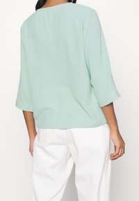 Haut léger vert menthe, coupe décontractée, encolure bateau flatteuse et manches trois-quarts. Associé à un pantalon blanc, visible de dos.