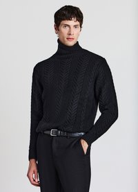 Schwarzer Strickpullover mit hohem Kragen und Fischgrätmuster, kombiniert mit schwarzen Hosen und einem eleganten Gürtel.