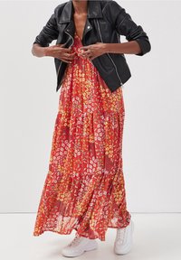 Robe maxi rouge à motif floral avec un design en couches, ornée d'imprimés jaunes et blancs. Associée à une veste en cuir noire et des baskets blanches.